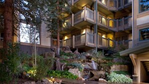 A multi-story hotel with balconies, a landscaped garden with a small waterfall, and a stone pathway. - Warm and Cozy 3 Bedroom Ski In, Ski Out Condo in Snowmass Base Village (Snowmass Village)