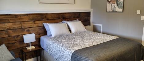 Un dormitorio con cabecera de madera, cama con ropa de cama estampada, mesita de noche con lámpara y un sofá.