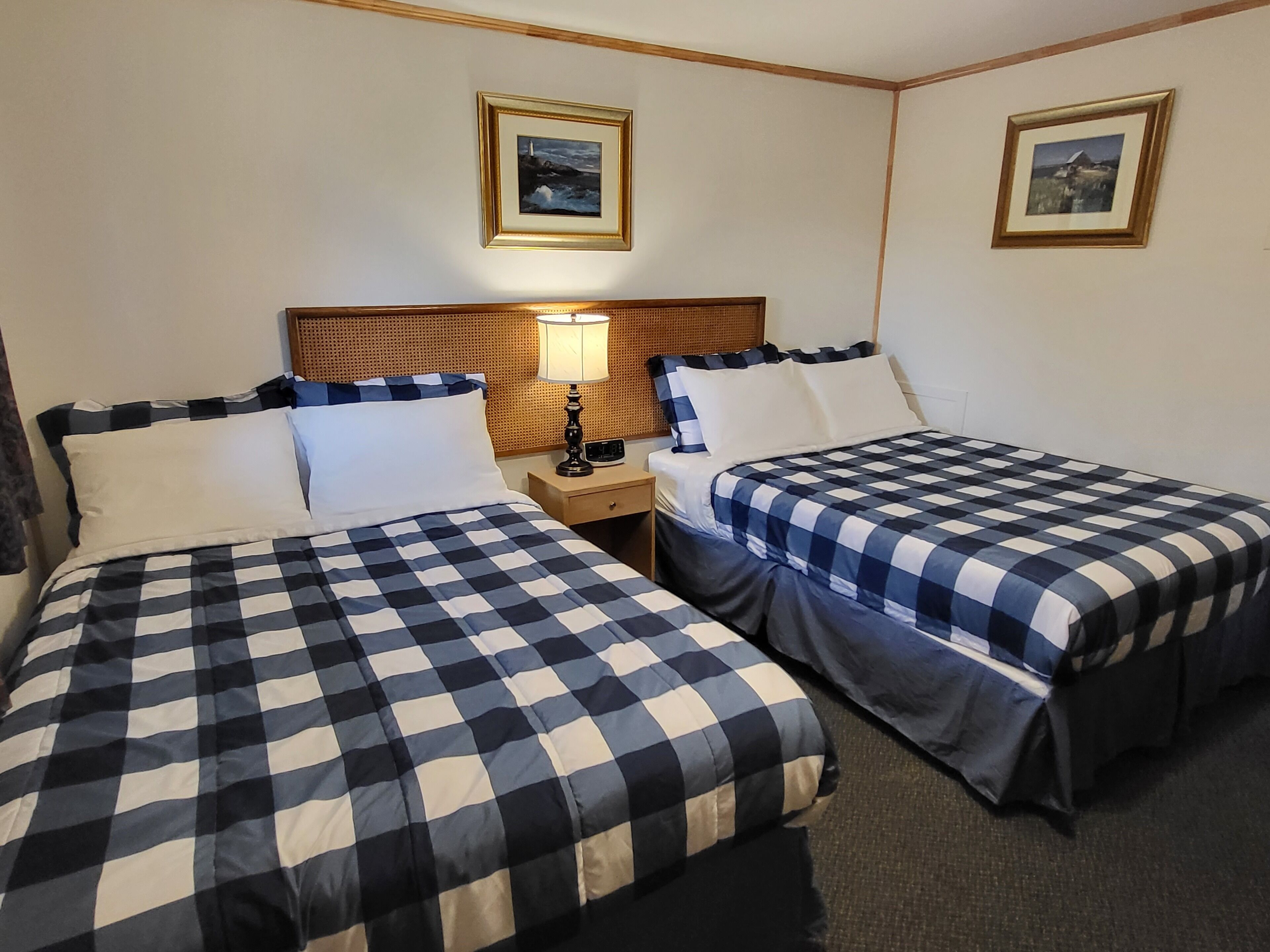 Habitación de hotel con dos camas, cada una con cubrecama a cuadros, cabecero de madera, mesita de noche con lámpara y dos cuadros enmarcados en la pared.