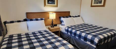 Habitación de hotel con dos camas, cada una con cubrecama a cuadros, cabecero de madera, mesita de noche con lámpara y dos cuadros enmarcados en la pared.