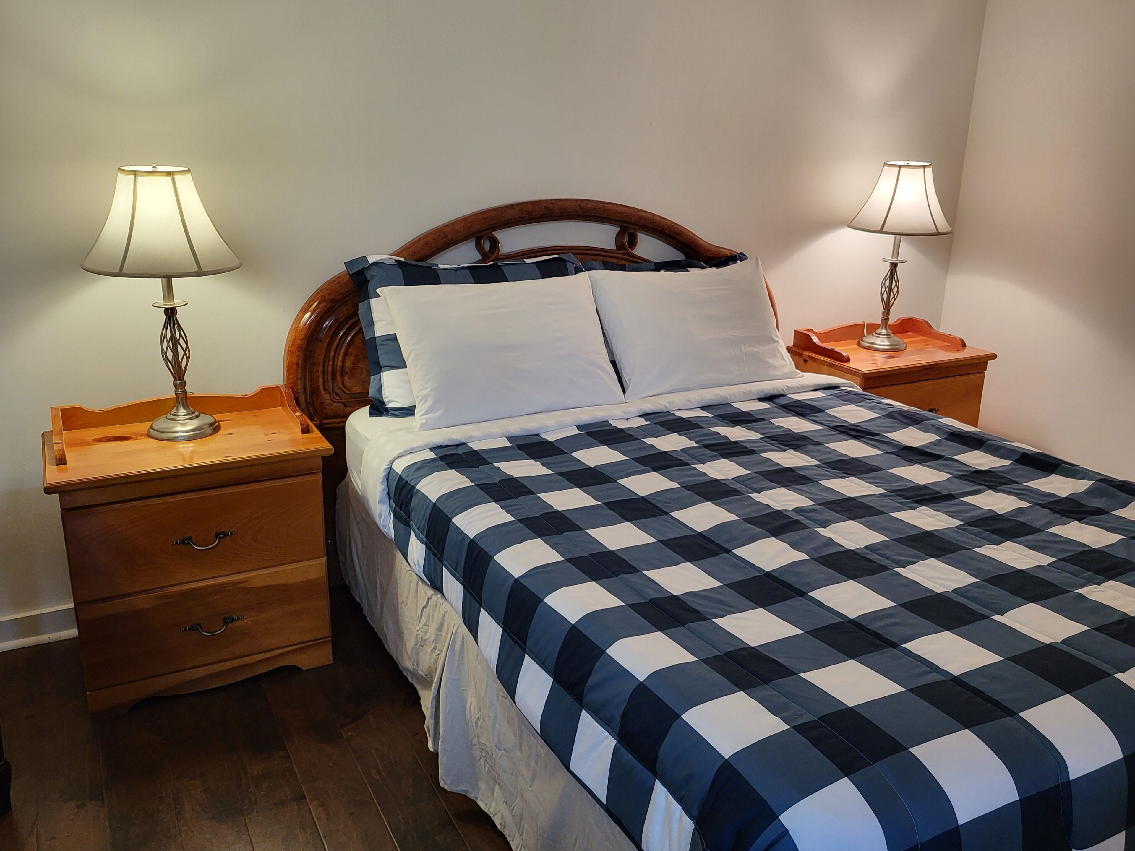 A neatly made bed with a checkered bedspread, two bedside lamps, and a wooden nightstand with a drawer.
