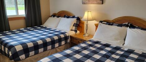A hotel room with two beds, checkered bedding, a nightstand with a lamp, a painting on the wall, and a window with curtains.