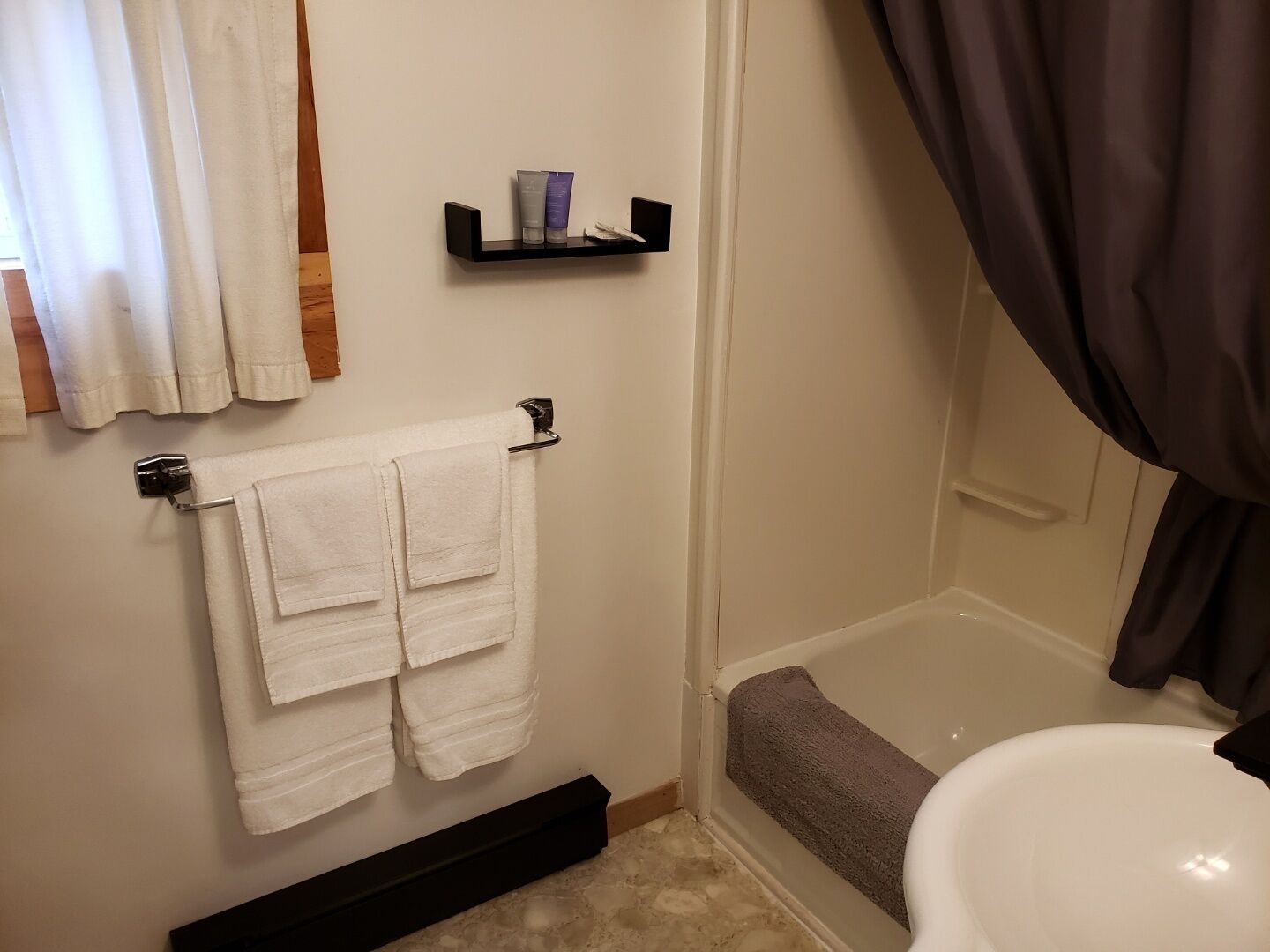 A bathroom with a white bathtub, a white toilet, and a towel rack with two white towels. There is a shelf with toiletries above the towel rack.