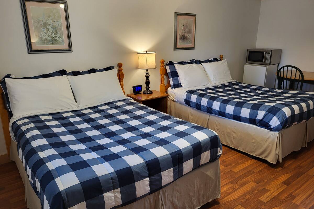 Habitació d'hotel amb dos llits, terres de fusta, una tauleta de nit amb una làmpada, un rellotge i un microones.