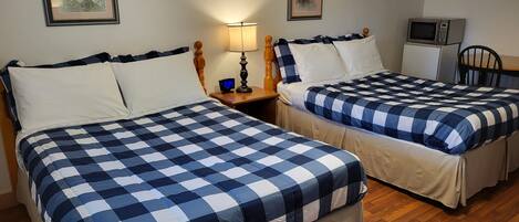A hotel room with two beds, wooden floors, a nightstand with a lamp, a clock, and a microwave.