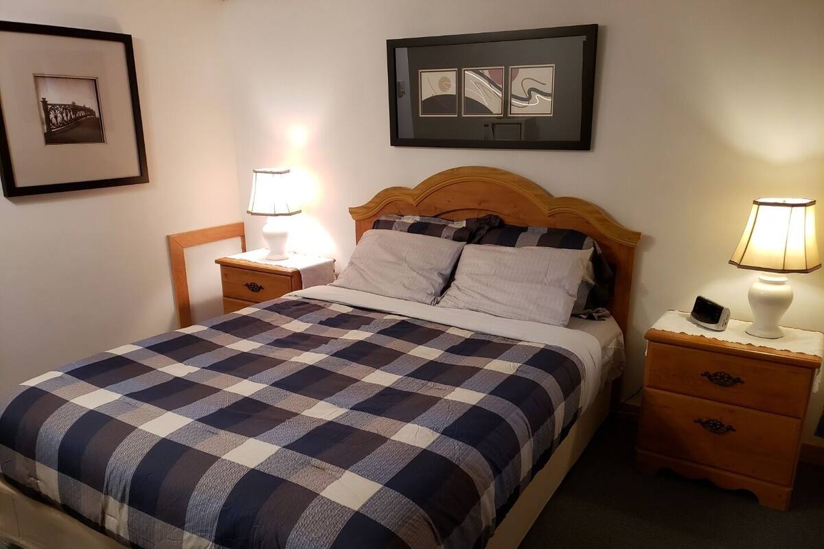 Un llit ben arrenjat amb un llençol estampat de quadradets, dues taules de nit amb làmpades i quadres a les parets.