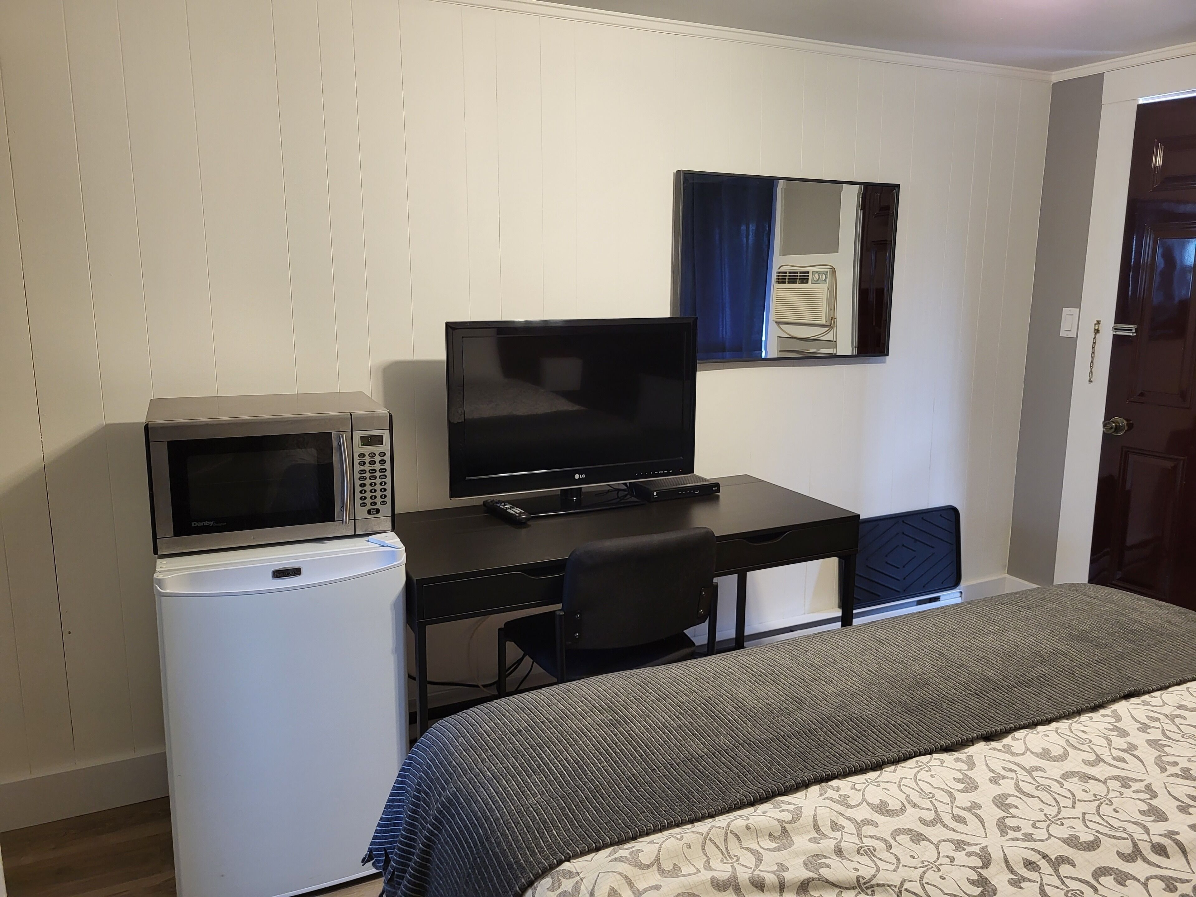 Ein kompaktes Hotelzimmer mit Küchenzeile, Flachbildfernseher, kleinem Kühlschrank, Schreibtisch und Bett mit gemustertem Überwurf.