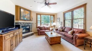 A living room with a fireplace, a television, a ceiling fan, and large windows. - Red Hawk Townhomes 2 Bedroom Rental in River Run Village (Keystone)