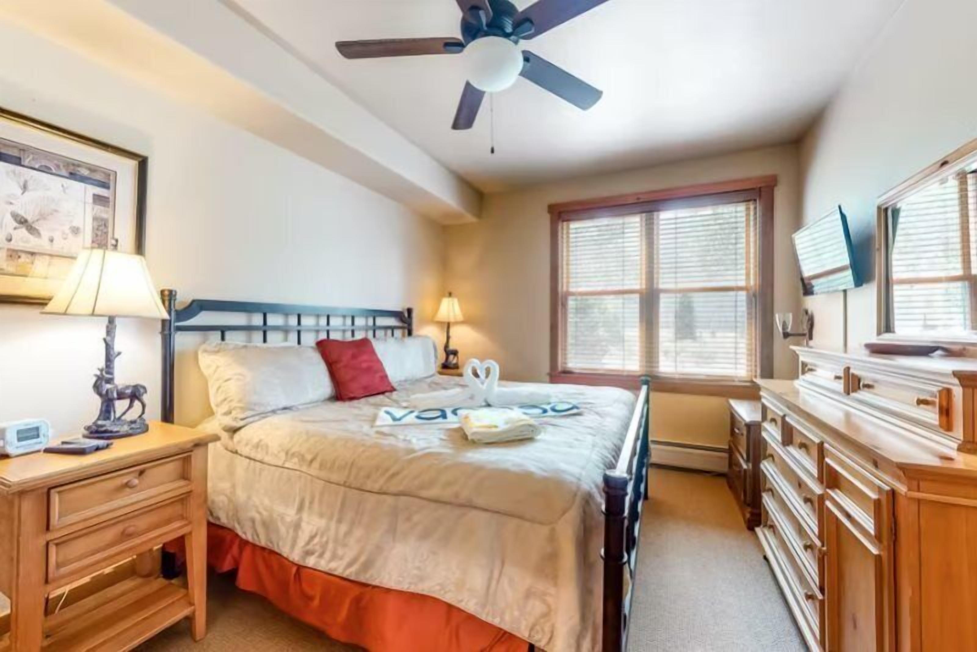 Ein Schlafzimmer mit einem Bett, Nachttischlampen, einem Deckenventilator, einem Fenster mit Jalousien, einem Schminktisch und einem gerahmten Bild an der Wand.