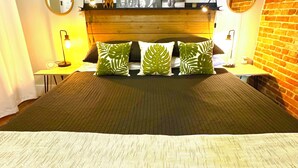 Una cama con cabecera de madera, almohadas con estampado de hojas verdes, una colcha negra y un cuadro colgado en la pared sobre la cama.