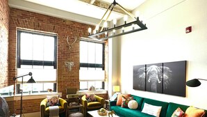 Una sala de estar acogedora con un sofá verde, un sillón amarillo y una mesa de centro blanca. Se observa una pared de ladrillo y un ventanal con persianas.