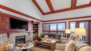A living room with a stone fireplace, wooden ceiling beams, a large flat-screen TV, and a cozy seating area with a sofa and armchairs. - Exclusive 4 Bedroom Ski in, Ski out Vacation Rental With Hot Tubs and Heated Outdoor Pool in Lionshead Village (Vail)