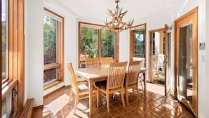 Un comedor con mesa y sillas de madera, ventanas amplias y una araña.