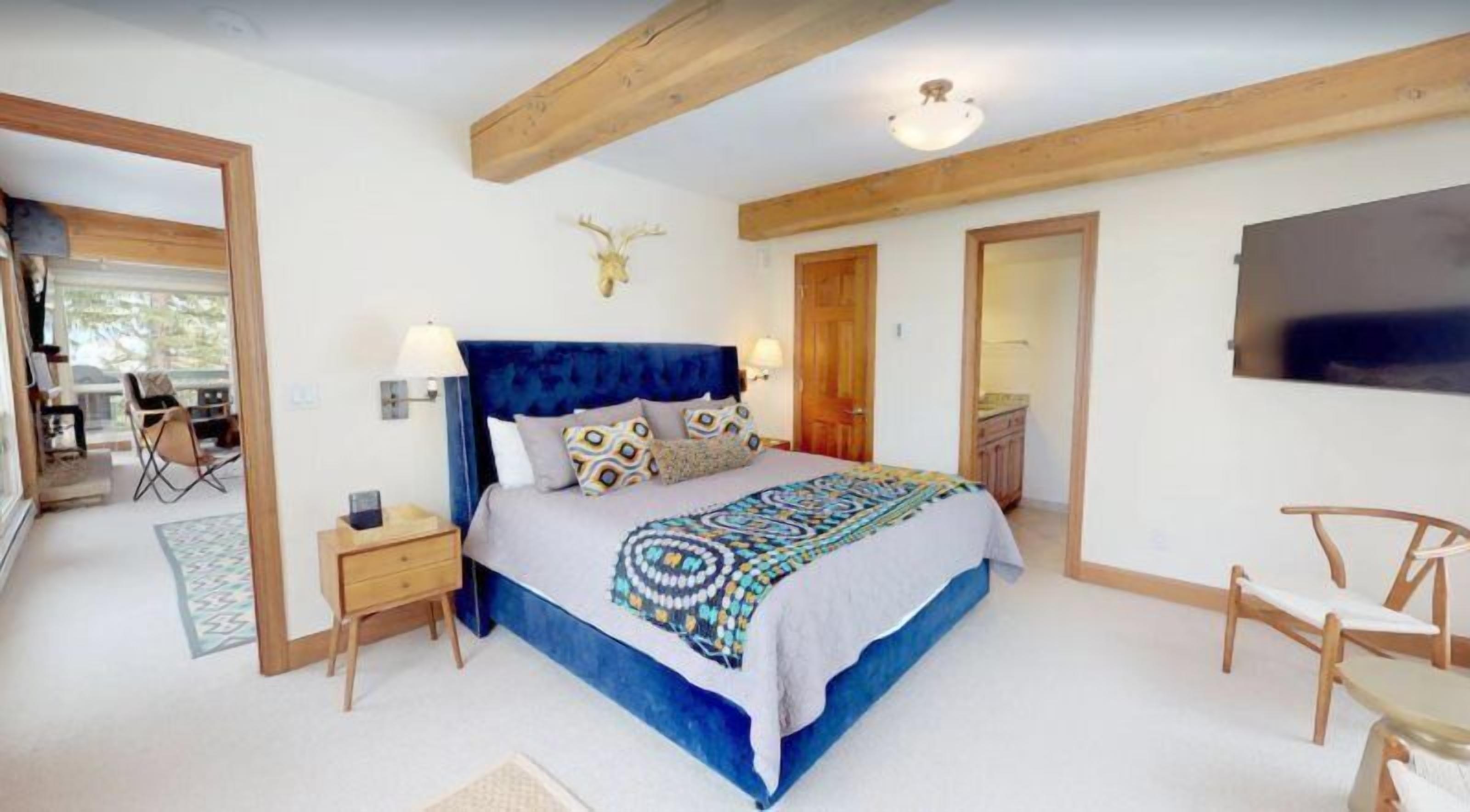 A bedroom with a large bed, wooden headboard, and a ceiling fan. There is a wooden door leading to another room and a small table with chairs.