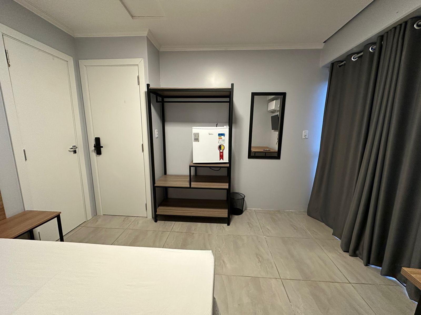 A compact room with a bed, a small table, a wardrobe, a mirror, and a window with curtains.