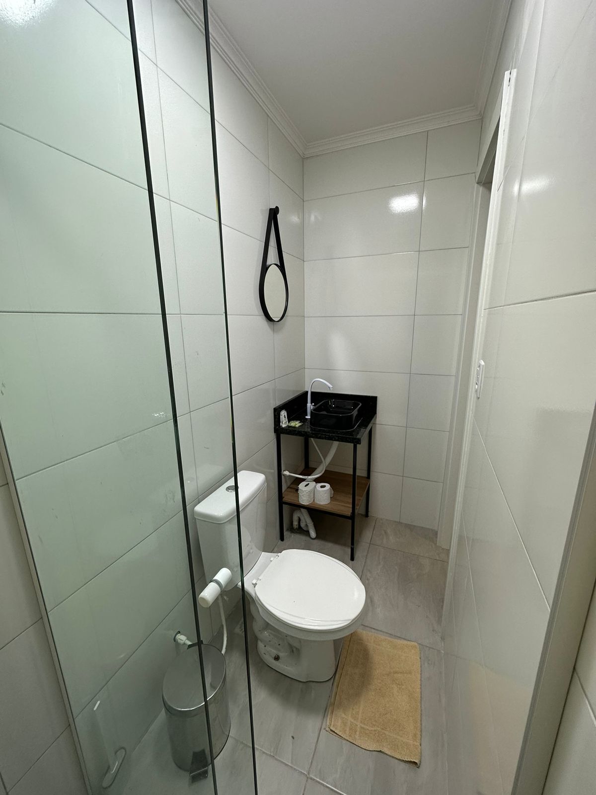 Un baño pequeño y funcional con inodoro, cabina de ducha de vidrio y un mueble negro con lavamanos y toallero.