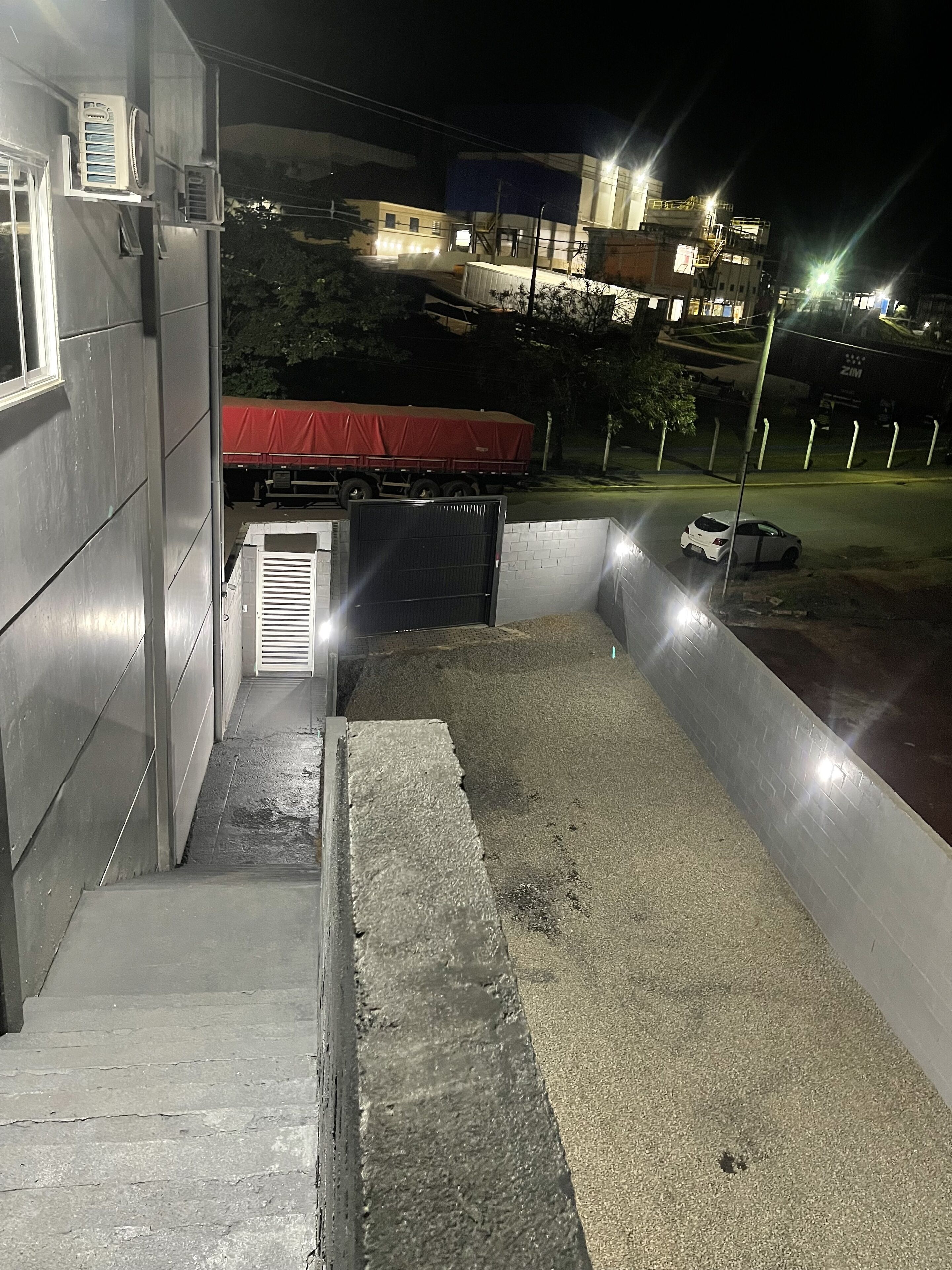 A nighttime view of a building with a gated entrance, a red-covered vehicle parked inside, and a white car parked outside.