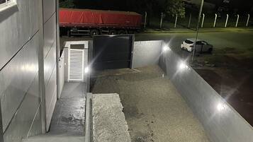 A nighttime view of a building with a gated entrance, a red-covered vehicle parked inside, and a white car parked outside.