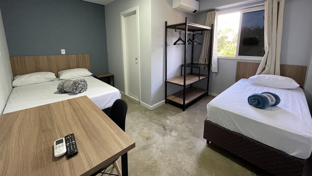 Habitación de hotel compacta con dos camas, una mesita con un control remoto, una cama litera con un perchero y una ventana con cortinas.