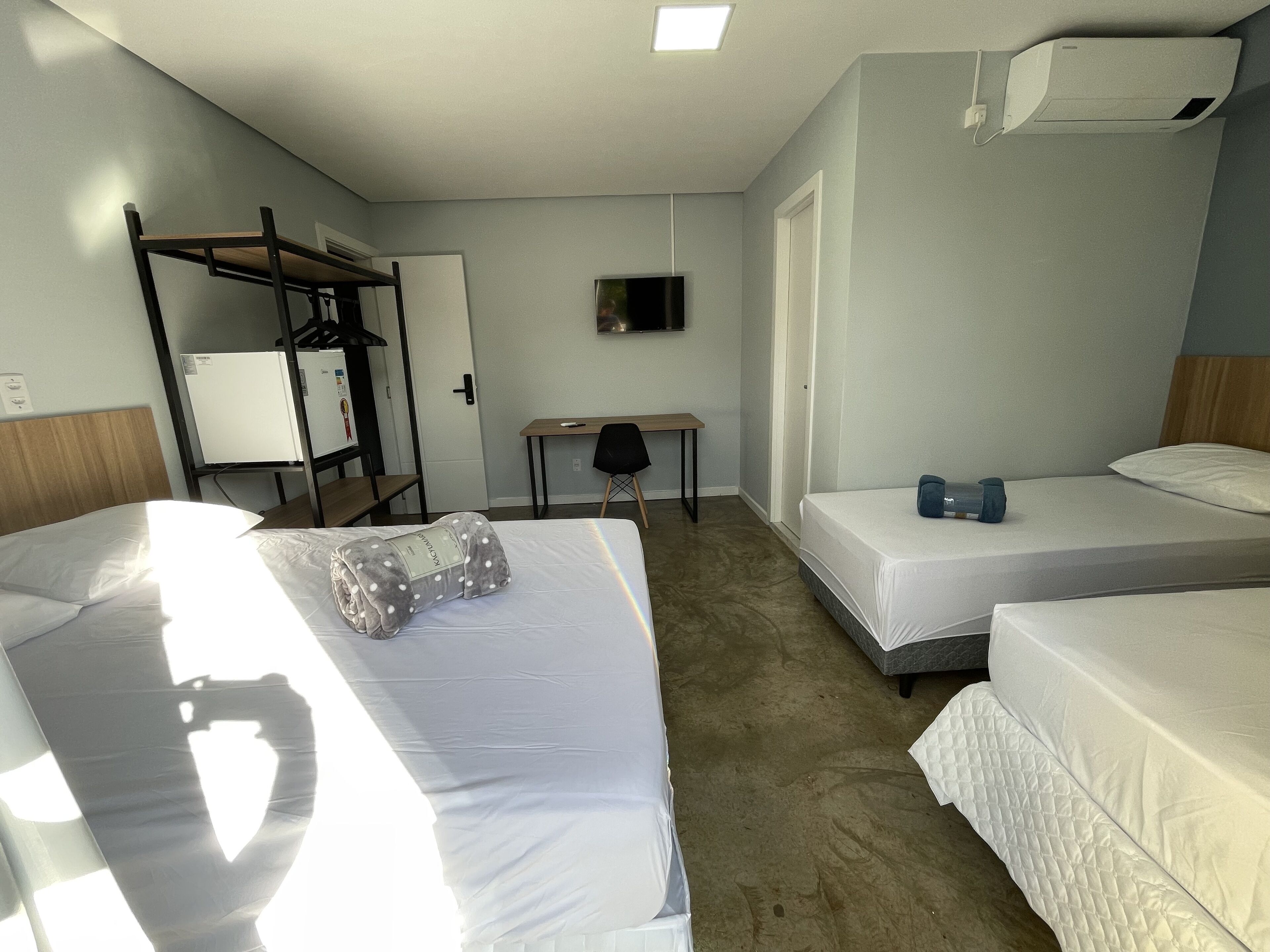 A hotel room with three beds, a desk, a chair, a TV, and an air conditioning unit.