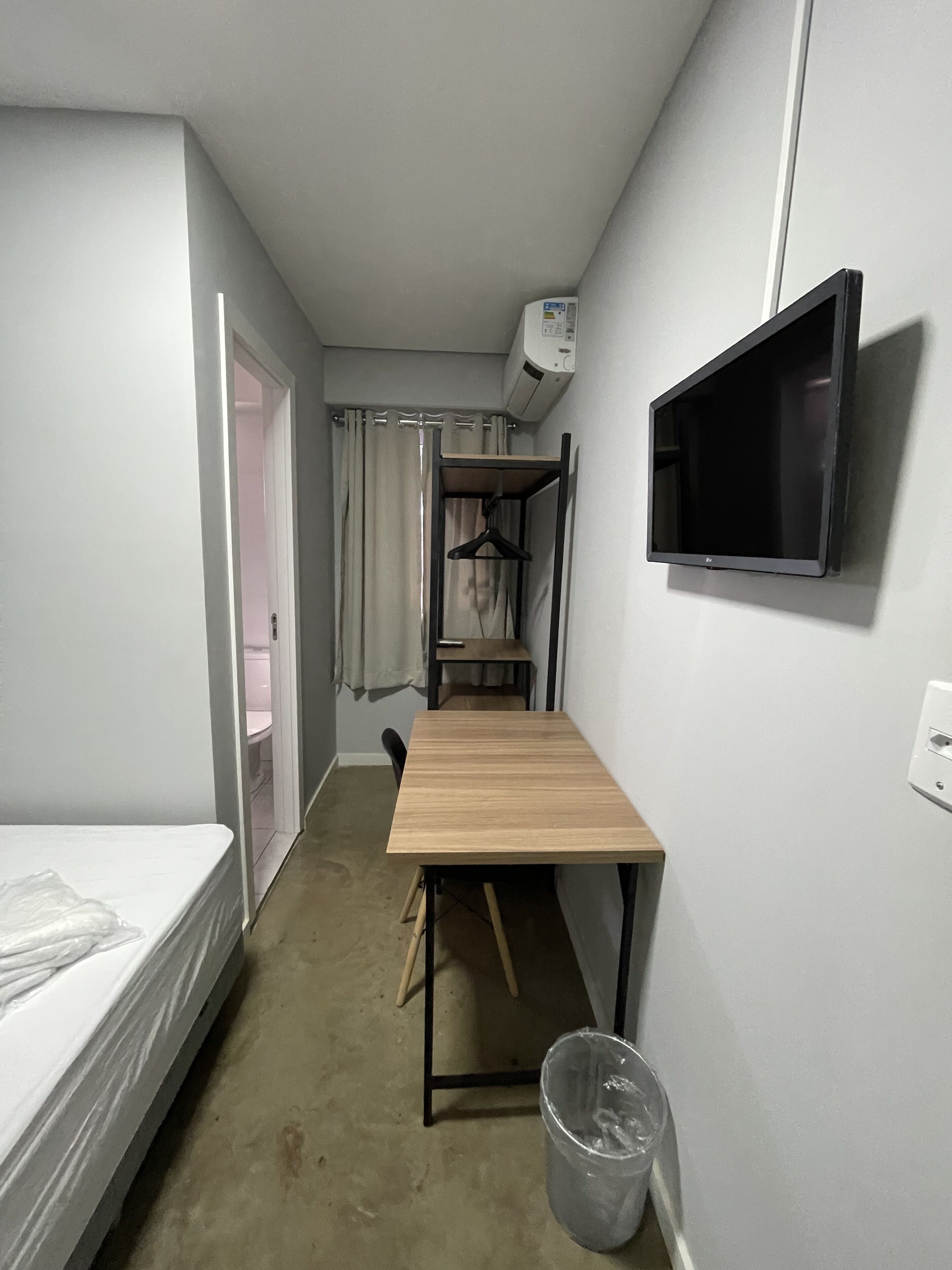 A compact hotel room with a single bed, a small wooden table, a TV mounted on the wall, and a closet with a hanging rack.