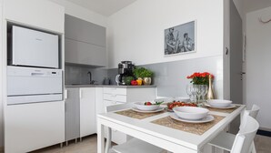 A modern kitchen with white cabinetry, a built-in oven, and a coffee maker. There is a dining table set with white plates and a vase of red flowers. - Seaside Apartamenty - Dominikana (Ustronie Morskie)