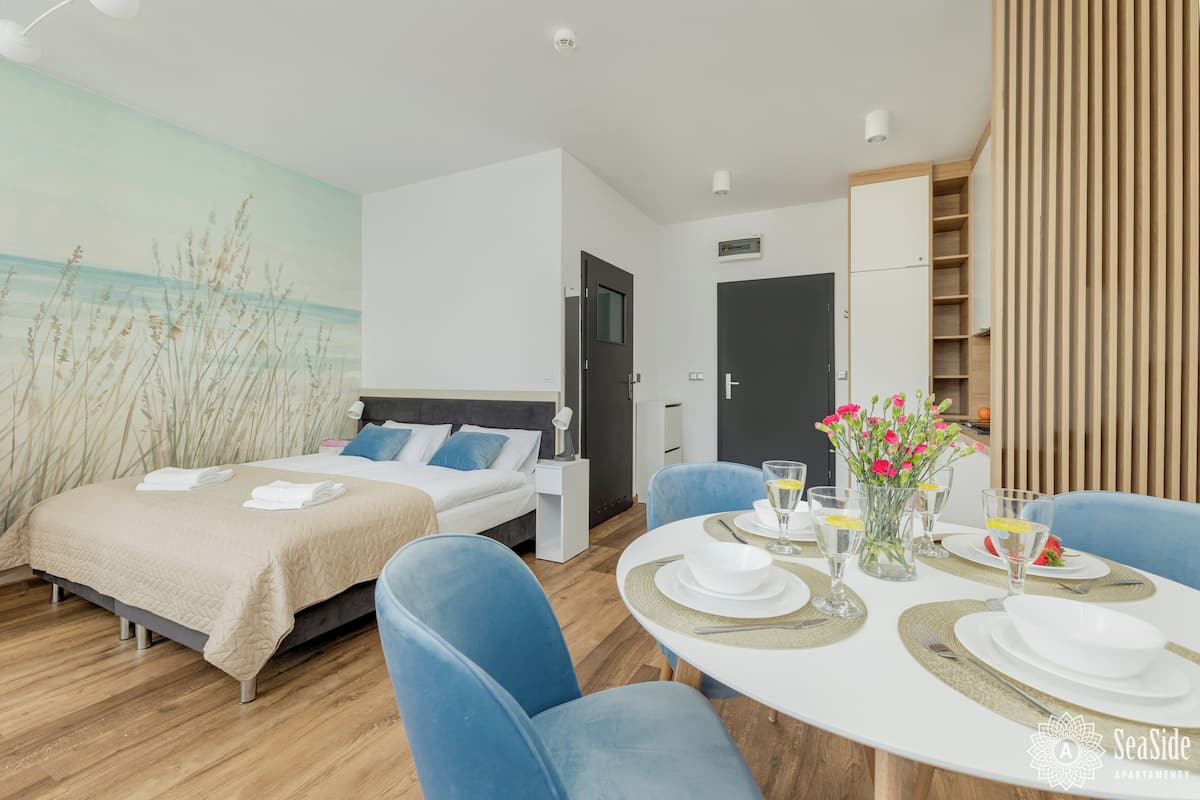 Una habitación de hotel moderna con una cama, dos sillas azules, una mesa de comedor con platos y vasos, y una pared decorada con un mural de playa.