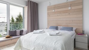 A modern bedroom with a large bed, a wooden headboard, a window with a view of trees, and a bedside table with a lamp. - Seaside Apartamenty - Dominikana (Ustronie Morskie)