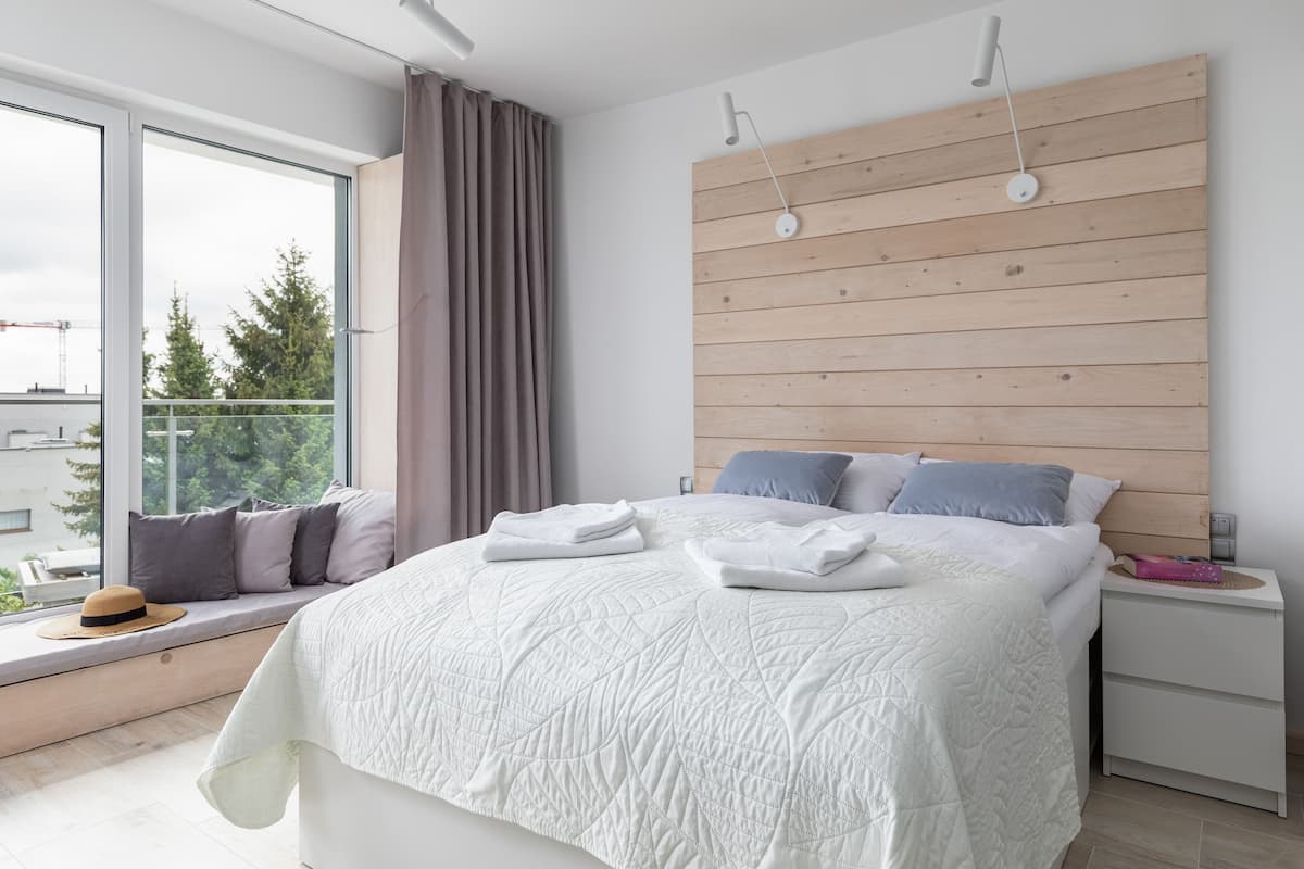 Un dormitorio moderno con una cama grande, cabecera de madera, ventana con vistas a árboles y una mesita de noche con lámpara.