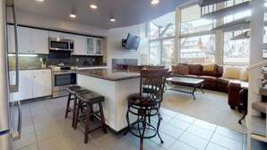 A modern kitchen with a central island, bar stools, a dining area with a sofa and coffee table, and a large window offering a view of buildings. - 1 Bedroom Boutique Resort Condo With Hot Tub Access and Within Walking Distance to the Eagle Bahn Gondola (Vail)