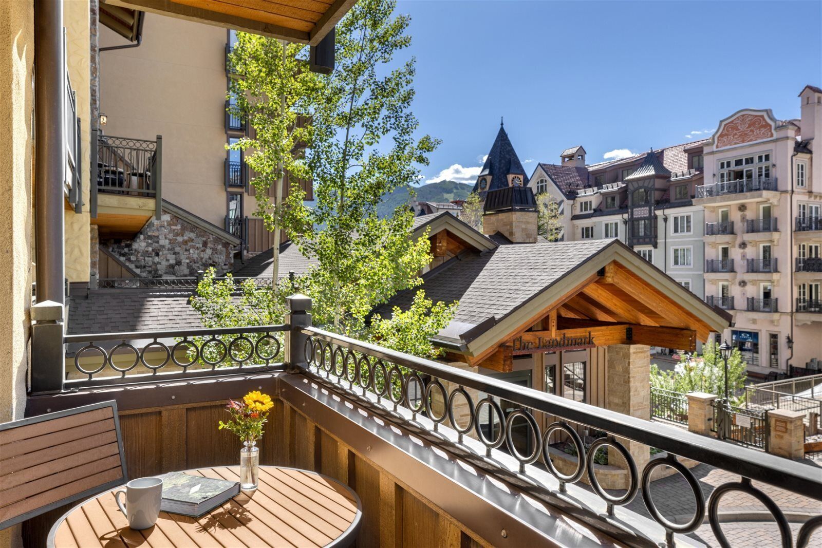 A balcony with a wooden table and a vase of flowers, overlooking a town with historic buildings and a mountain in the background.