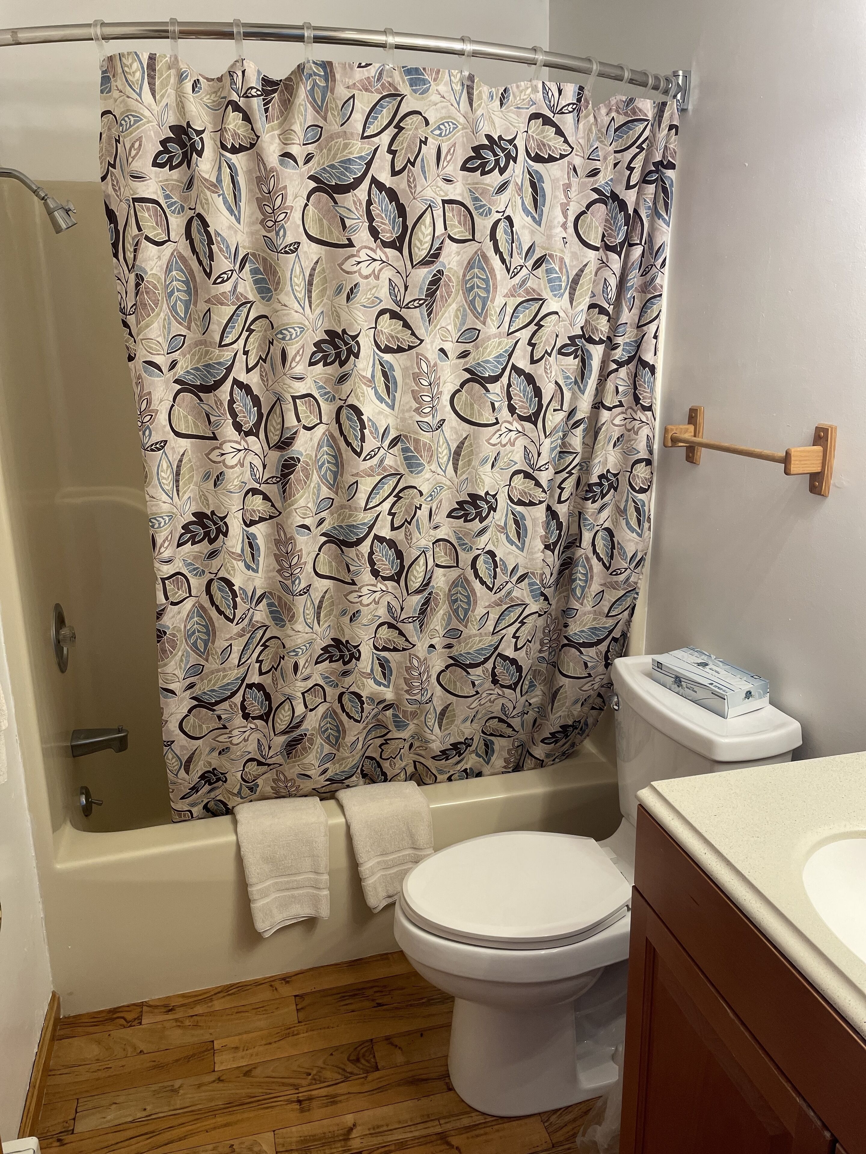 A bathroom with a shower curtain featuring a leaf pattern, a white toilet, and a wooden floor.