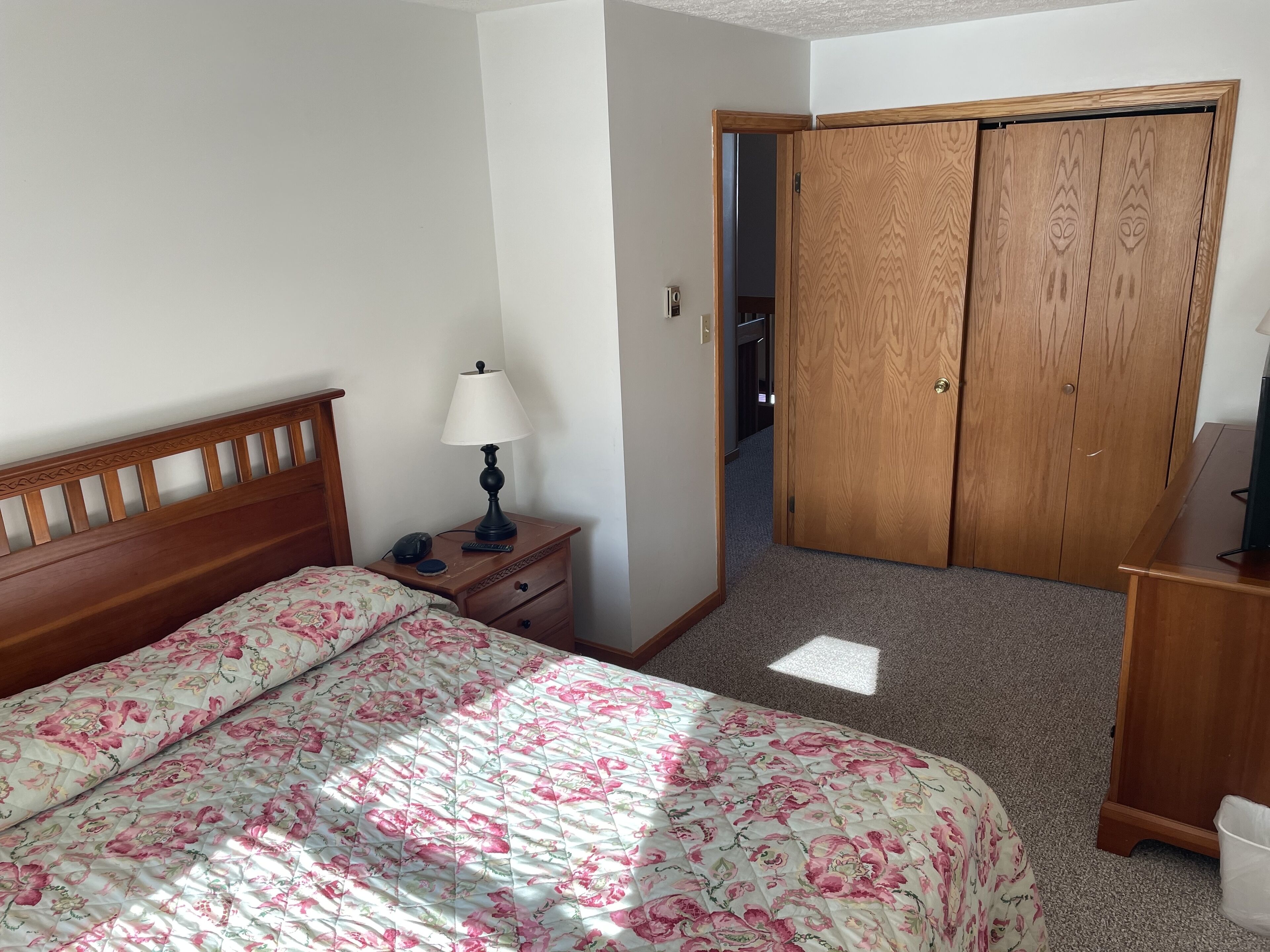 A bedroom with a wooden bed, a nightstand with a lamp, a dresser, and a closet.