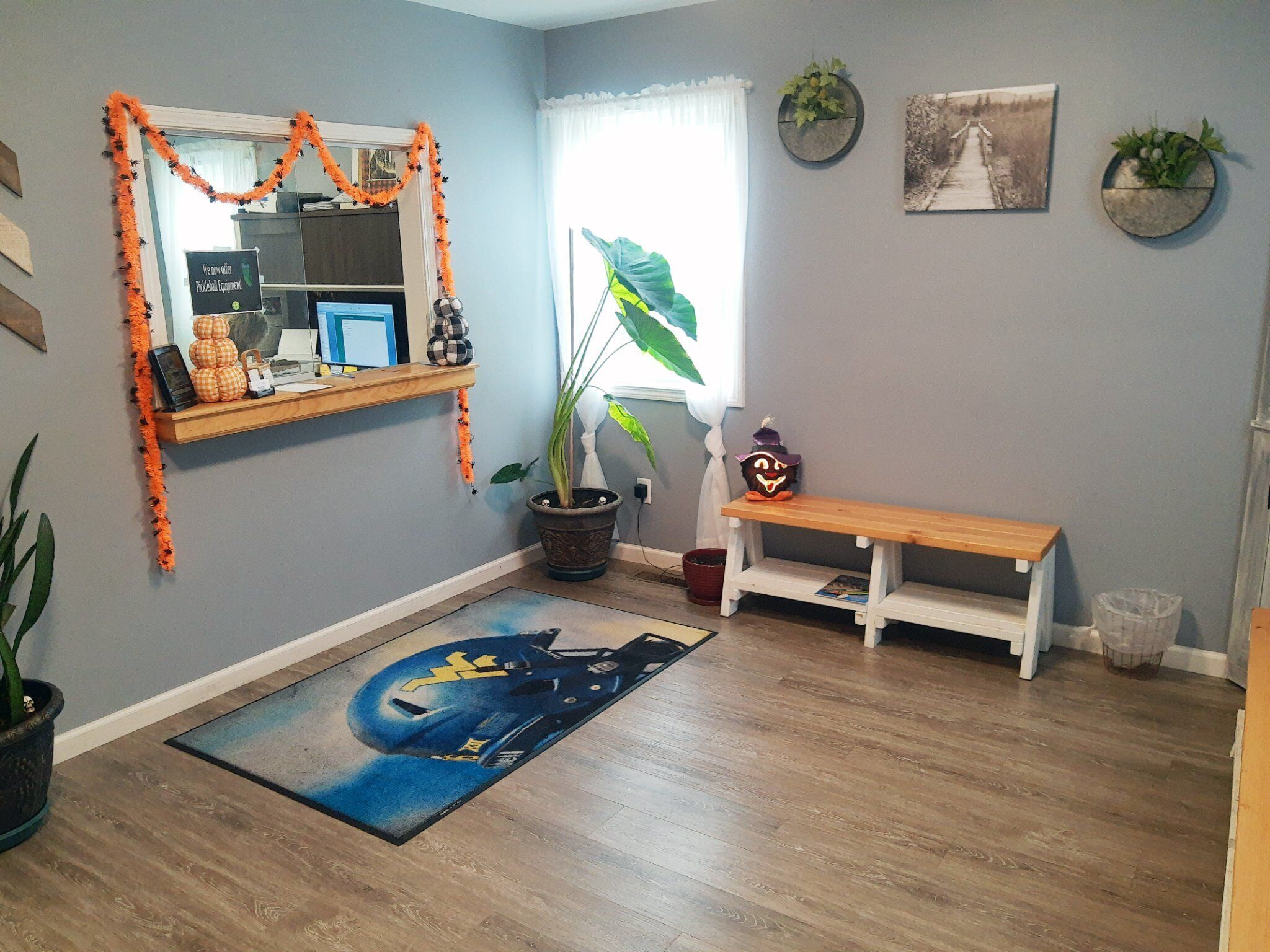 A room with a wooden floor, a blue mat with a car design, a bench, a potted plant, a mirror with orange decorations, and a sign on the wall.