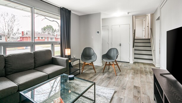 Un salon moderne avec un canapé gris, deux fauteuils noirs, une table basse en verre et un téléviseur posé sur un meuble en bois.