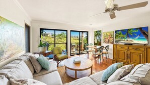 A spacious living room with a large window, a ceiling fan, and a television. - Poipu Beach Kauai Honeymoon: View Sea Turtles From Your Oceanfront Deck-AC (Koloa)