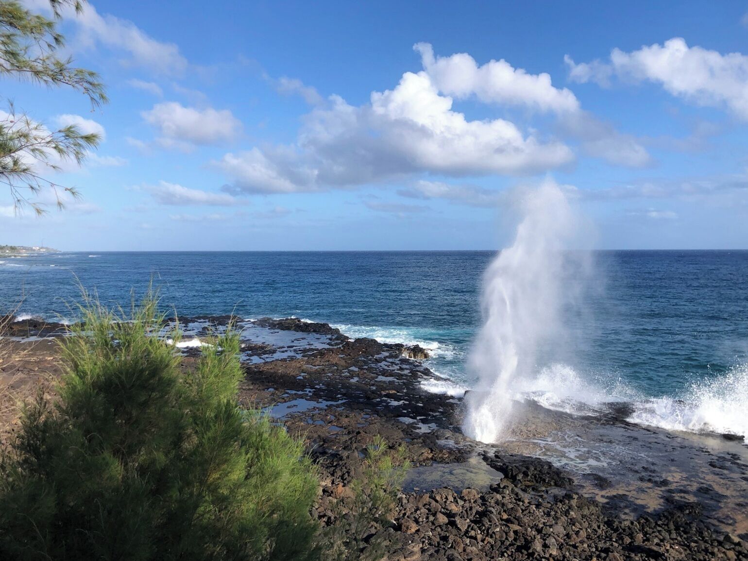 A coastal landscape with a rocky shoreline, a clear sky, and a visible water spout.