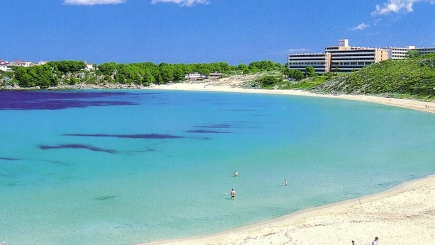 A beach with clear turquoise water, a sandy shore, and a building in the background. - Lovely 2-bedrooms condo just 300 mts from beach (Arenal d`en Castell)