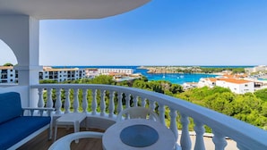 A balcony with a white balustrade, a blue sofa, a small round table, and a view of a coastal town with buildings and a blue sea. - Lovely 2-bedrooms condo just 300 mts from beach (Arenal d`en Castell)