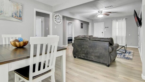 Un salon moderne avec un canapé gris, des chaises de salle à manger blanches, une table de salle à manger en bois, un ventilateur de plafond et un téléviseur à écran plat fixé au mur.