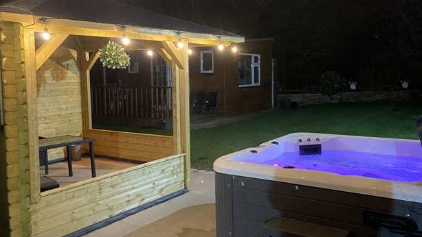 A hot tub with a wooden deck and string lights, a covered seating area, and a lawn in the background. - The Heights & Hollows Farmhouse (Downpatrick)