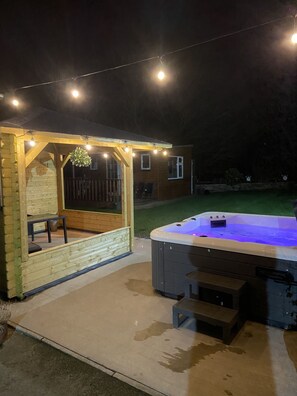A hot tub with a wooden deck and string lights, a covered seating area, and a lawn in the background. - The Heights & Hollows Farmhouse (Downpatrick)