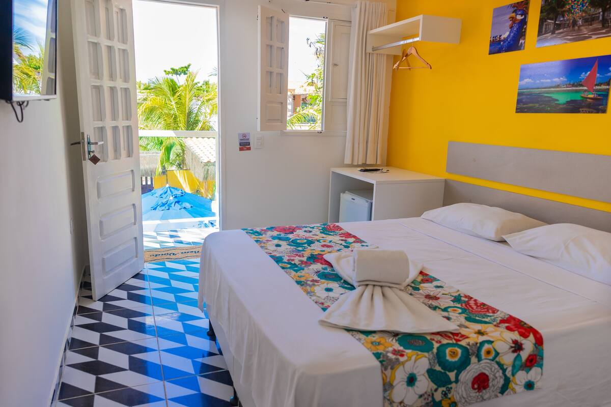 Une chambre d’hôtel avec un lit, une petite table de chevet, une fenêtre avec des rideaux, un mur décoré de tableaux et une porte donnant sur l’extérieur.