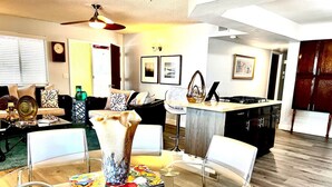 Una sala de estar moderna con una mesa de comedor de vidrio, sillas blancas y una zona de cocina al fondo.