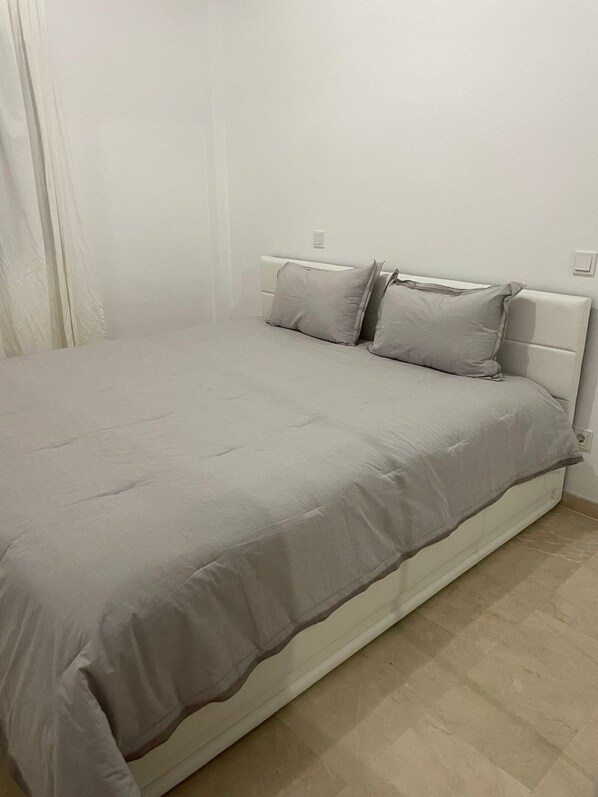 Un lit avec une couvre-lit et des oreillers gris, une tête de lit blanche et un plancher de couleur claire.