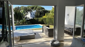 A modern living room with a glass door leading to a pool area, outdoor furniture, and a view of a white building and greenery. - Luxurious sea view villa for 6 people, 1km from the beach (Marbella)