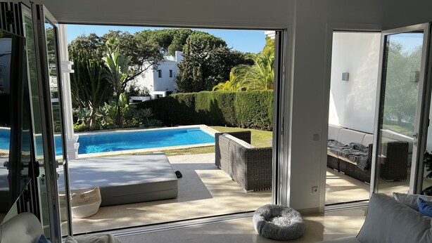 A modern living room with a glass door leading to a pool area, outdoor furniture, and a view of a white building and greenery. - Luxurious sea view villa for 6 people, 1km from the beach (Marbella)