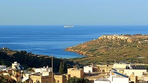 A coastal town with buildings, a ship in the distance, and a rocky hill. - Penthouse with Private Roof Top Pool (Ix-Xaghra)