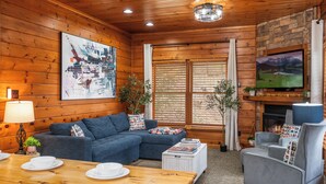 Interior acogedor de una cabaña con techo de madera, un cuadro grande, chimenea, televisión y zona de comedor con una mesa dispuesta para dos.
