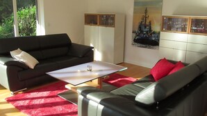 A modern living room with a black leather sofa, a red rug, and a glass coffee table. There is a white cabinet with glass doors and a large cityscape painting on the wall.